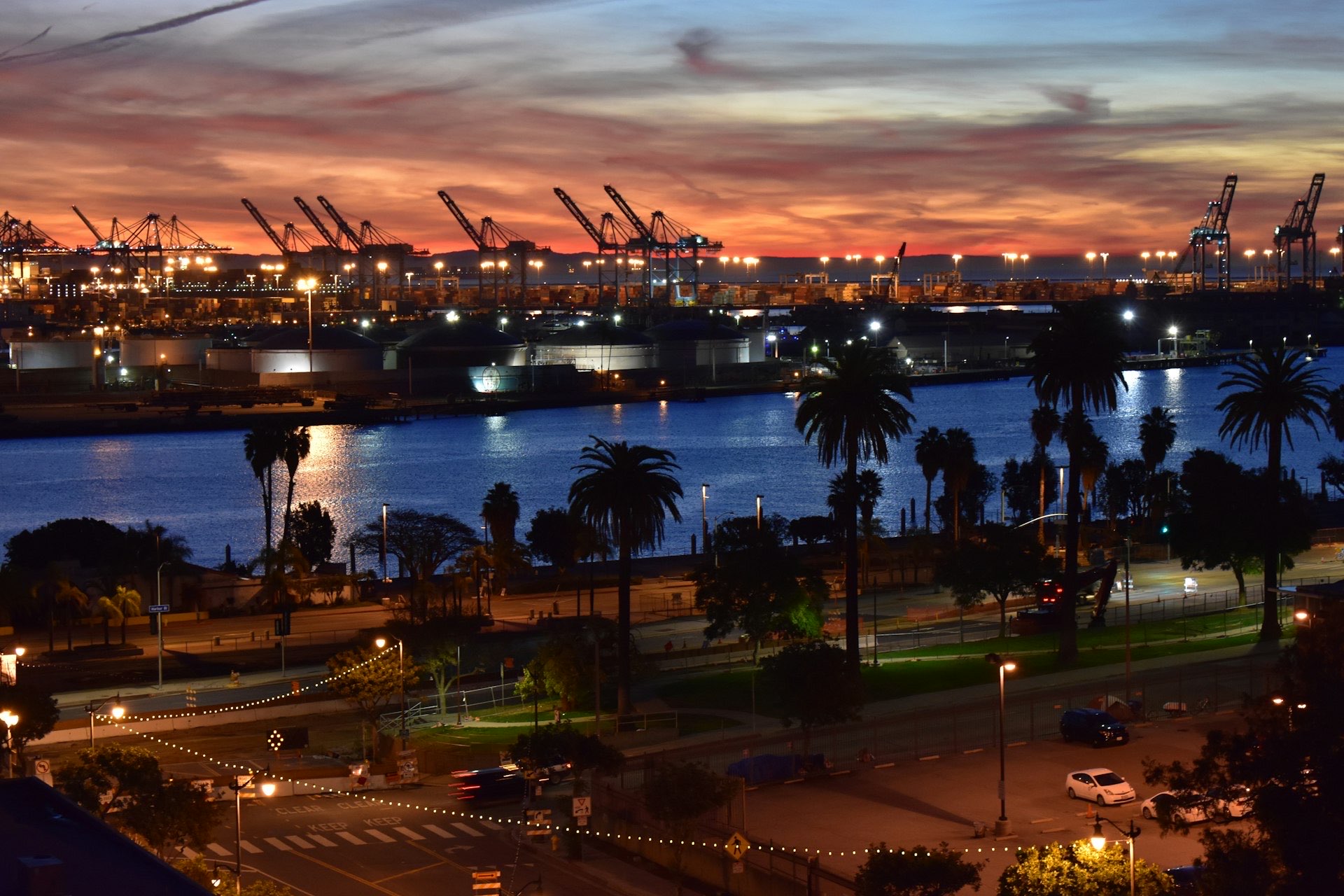 Container cranes and palm trees silhouetted against an ember-orange sunset over the Los Angeles–Long Beach harbor