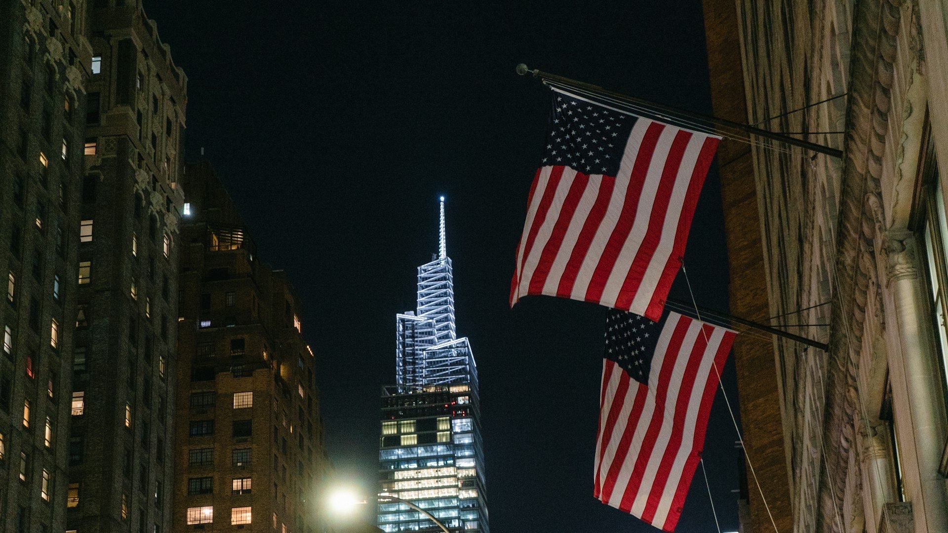 Two American flags on angled poles between dark masonry buildings at night, a lit tower behind