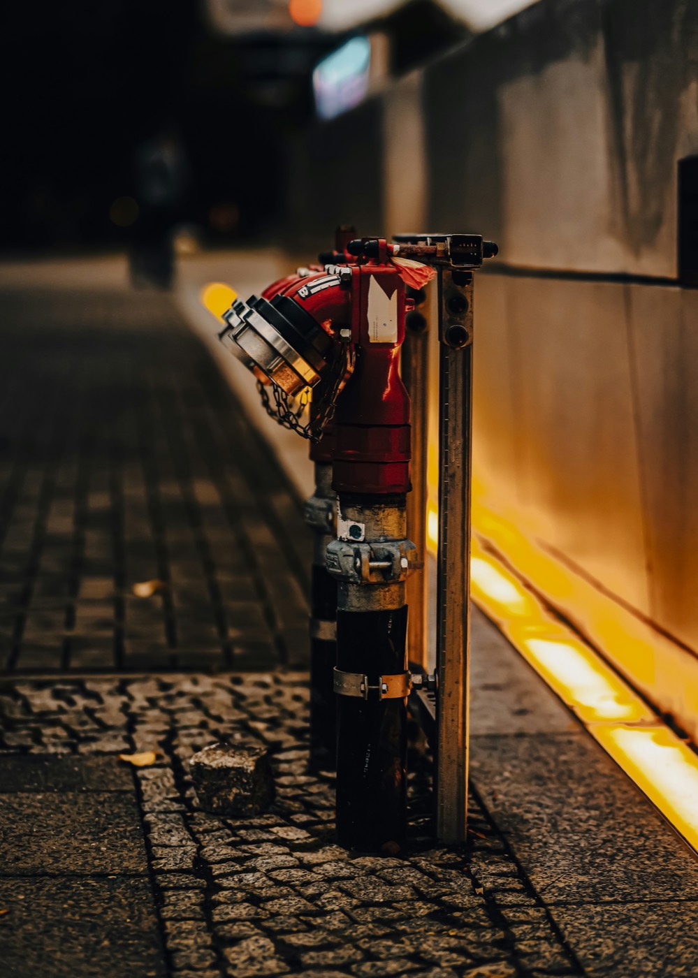 Sidewalk standpipe connection at night beside a wall lit by warm amber strip lighting
