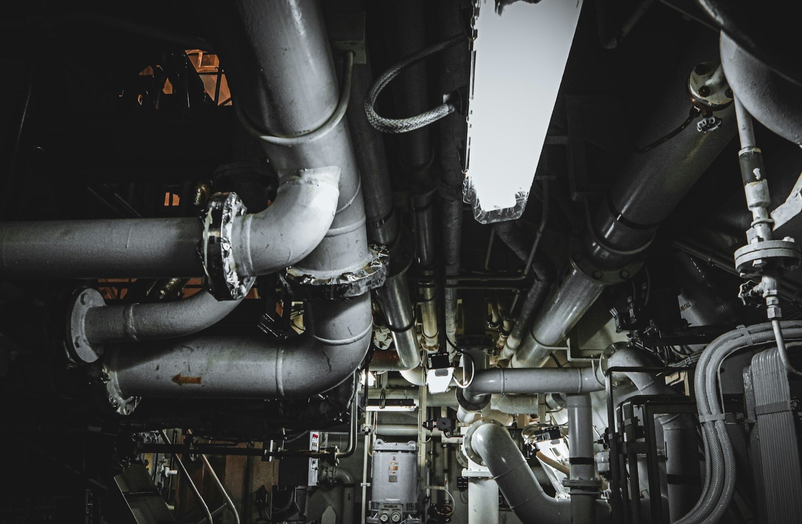 Dense overhead network of gray industrial pipes and valves in a dark machine room