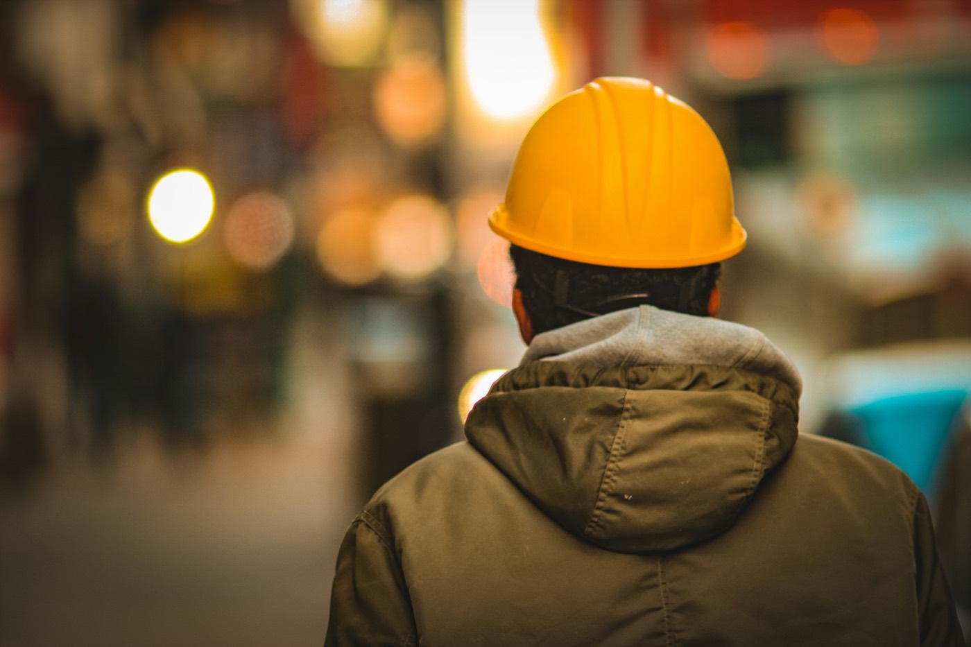 Worker in a yellow hard hat and hooded jacket seen from behind against warm night lights